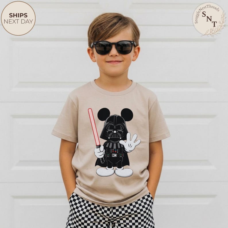 May include: Tan short-sleeved t-shirt featuring a cartoon Darth Vader character with Mickey Mouse ears, holding a red lightsaber. The character is black and white. The shirt has the text "SnuggleNestThreads" and "SHIPS NEXT DAY" on it.