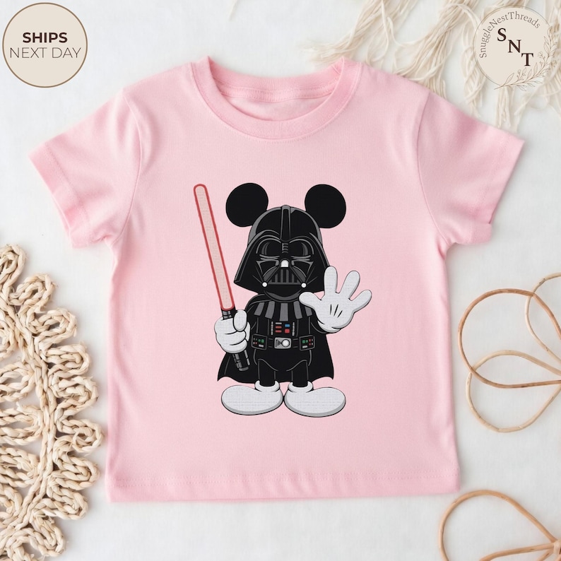 May include: A light pink t-shirt featuring a cartoon character mashup of Darth Vader and Mickey Mouse. The character is in black and white with a red lightsaber. The shirt has the text "SHIPS NEXT DAY" in a circle.