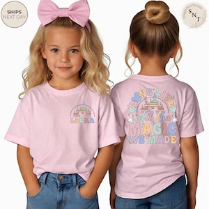 May include: Two young girls wearing light pink t-shirts with colorful graphics. One shirt features a castle design with the name "LAURA". The other shirt has the text "All the MAGIC WE MADE" with a castle illustration.