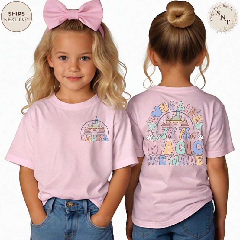 May include: Two young girls wearing light pink t-shirts with colorful graphics. One shirt features a castle design with the name "LAURA". The other shirt has the text "All the MAGIC WE MADE" with a castle illustration.