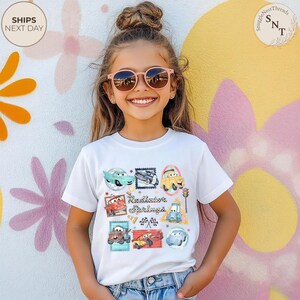 May include: A white t-shirt featuring a colorful graphic of various cartoon cars in postage stamp designs, with the text "Radiator Springs". The shirt is worn by a young person wearing pink sunglasses.
