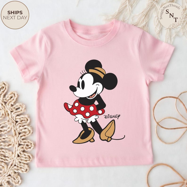 May include: A light pink t-shirt featuring a graphic of Minnie Mouse. Minnie is depicted in her classic outfit: a red skirt with white polka dots, yellow shoes, and a matching bow. The Disney logo is printed below the character.