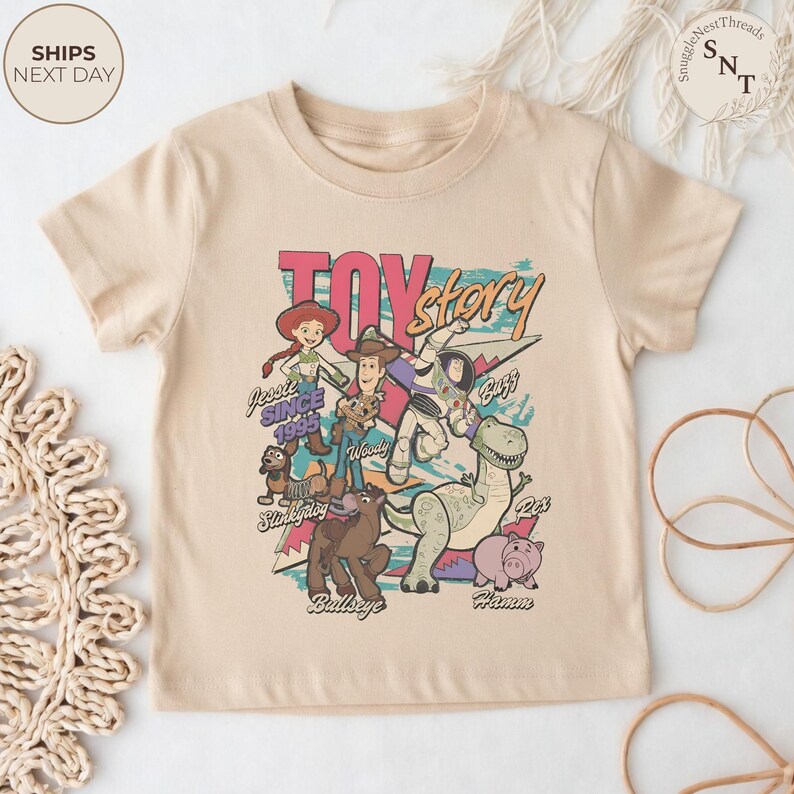 Puede incluir: Una camiseta beige de manga corta con un estampado gr&aacute;fico de los personajes de Toy Story, incluyendo a Jessie, Woody, Buzz Lightyear, Slinky Dog, Bullseye, Rex y Hamm. El texto dice Toy Story y Jessie Since 1995 con los nombres de los personajes.