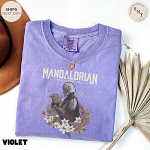 Puede incluir: Una camiseta violeta de manga corta con un gr&aacute;fico del Mandaloriano y Grogu rodeados de flores. El texto dice "Mandalorian" y "This is the way". La camiseta tiene cuello redondo y un aspecto de tela lavada de estilo vintage.
