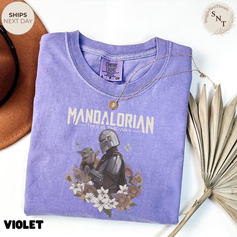 Puede incluir: Una camiseta violeta de manga corta con un gr&aacute;fico del Mandaloriano y Grogu rodeados de flores. El texto dice "Mandalorian" y "This is the way". La camiseta tiene cuello redondo y un aspecto de tela lavada de estilo vintage.