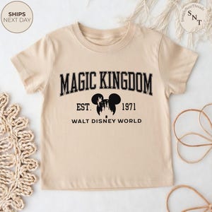 May include: A beige t-shirt featuring the text "MAGIC KINGDOM" in black, arched lettering. Below, it reads "EST. 1971 WALT DISNEY WORLD" with a Mickey Mouse silhouette. The shirt has short sleeves and a crew neck.