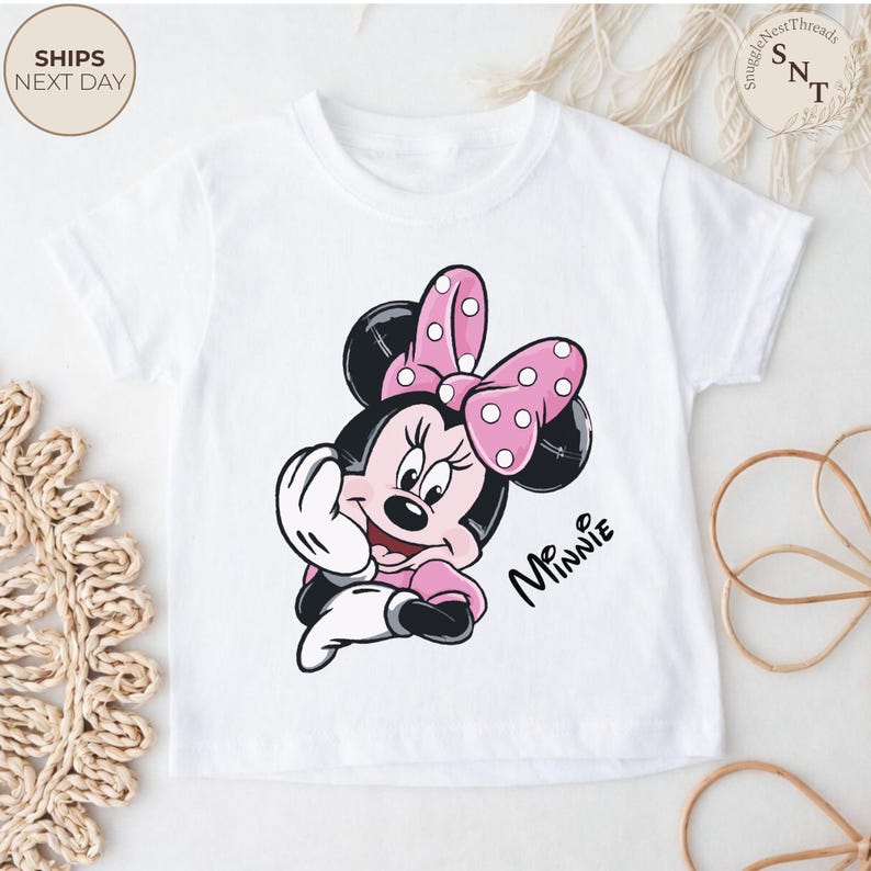 May include: White t-shirt featuring a cartoon illustration of Minnie Mouse. Minnie is depicted in pink and black with a pink polka dot bow. The word "Minnie" is written in black script. The shirt has short sleeves.