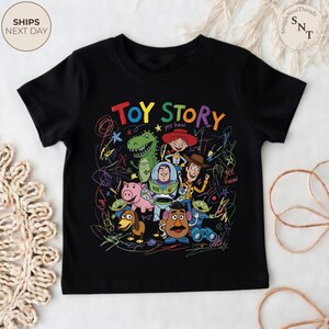 May include: Black t-shirt featuring a colorful graphic of characters from the animated film "Toy Story." The design includes the words "Toy Story" in large, multi-colored letters, along with images of Woody, Buzz Lightyear, and other characters. The shirt has a crew neck and short sleeves.