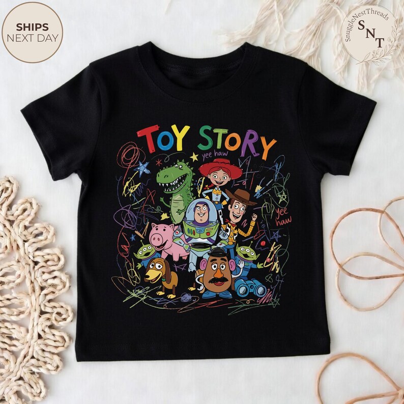 May include: Black t-shirt featuring a colorful graphic of characters from the animated film "Toy Story." The design includes the words "Toy Story" in large, multi-colored letters, along with images of Woody, Buzz Lightyear, and other characters. The shirt has a crew neck and short sleeves.