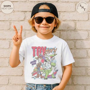 Może przedstawiać: Biała koszulka z kolorową grafiką "Toy Story" z Woodym, Buzzem i Rexem. Koszulka ma napis "TOY STORY" i "SINCE 1995". Dziecko ma na sobie czapkę z daszkiem i okulary przeciwsłoneczne.