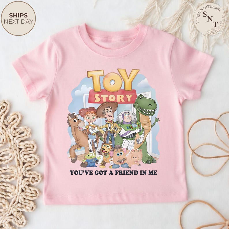 K&ouml;nnte beinhalten: Hellrosa T-Shirt mit einem Aufdruck von Toy Story-Charakteren und dem Schriftzug "TOY STORY" in Gold. Der Text "YOU'VE GOT A FRIEND IN ME" ist darunter gedruckt.