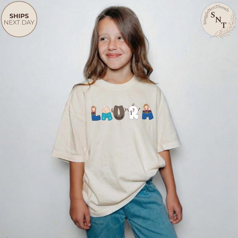 K&ouml;nnte beinhalten: Cremefarbenes T-Shirt mit dem Namen "LAURA" in einem verspielten, cartoonartigen Schriftzug auf der Vorderseite. Jeder Buchstabe ist mit einem anderen Charakter gestaltet. Das T-Shirt wird von einer Person getragen.