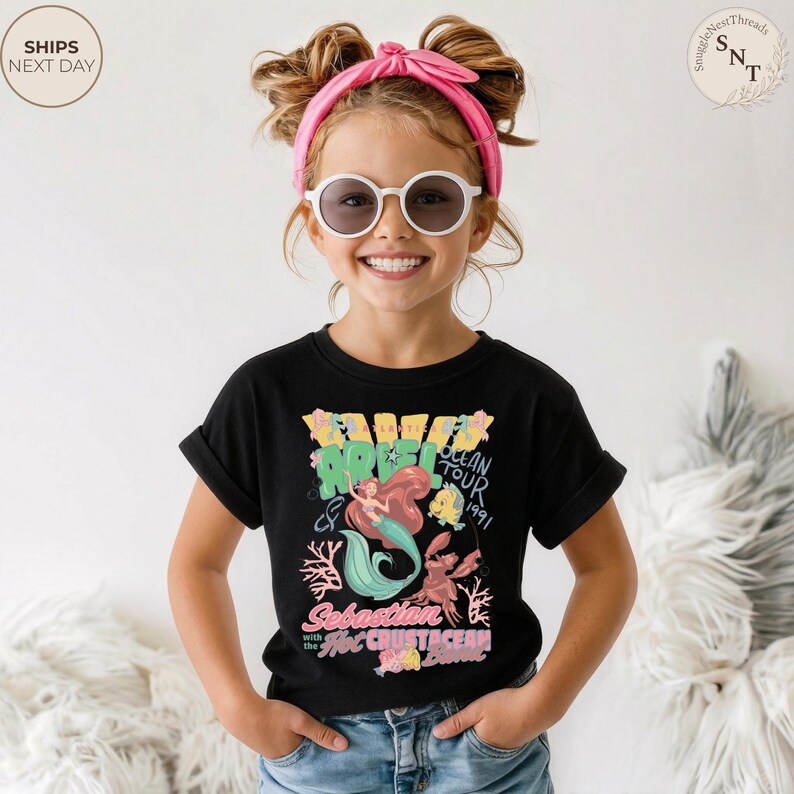 May include: A black short-sleeve t-shirt featuring a colorful graphic of Ariel the mermaid, Flounder, and Sebastian. The text reads Ariel Ocean Tour 1991, Sebastian and the Hot Crustacean Band. The shirt is paired with blue denim jeans.