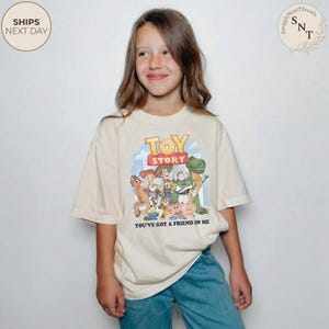 K&ouml;nnte beinhalten: Beiges T-Shirt mit farbenfroher Grafik von Figuren aus dem Animationsfilm "Toy Story". Das Shirt tr&auml;gt den Text "TOY STORY" und "YOU'VE GOT A FRIEND IN ME". Das T-Shirt wird von einer Person getragen.