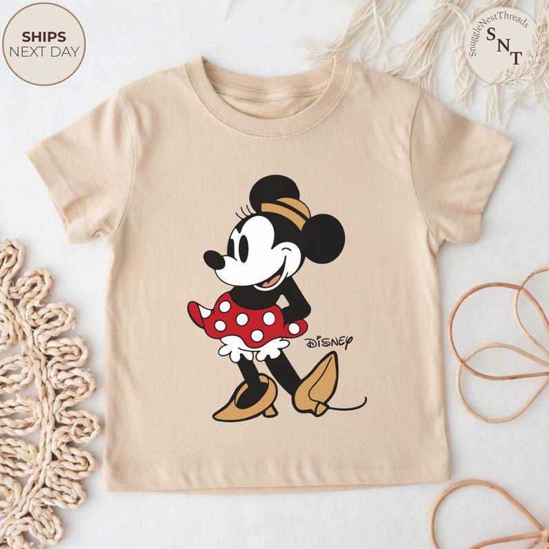 May include: A beige t-shirt featuring a graphic of Minnie Mouse in a red polka dot dress and yellow shoes. The Disney logo is printed on the shirt. The shirt has short sleeves.