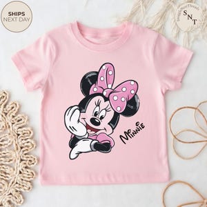 May include: A light pink t-shirt featuring a graphic of Minnie Mouse with a pink polka dot bow. The words "Mr. Minnie" are printed in black script. The shirt has short sleeves and a crew neck.