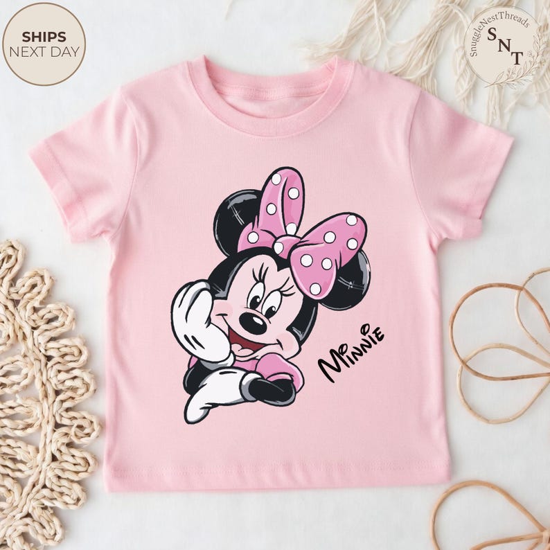 May include: A light pink t-shirt featuring a graphic of Minnie Mouse with a pink polka dot bow. The words "Mr. Minnie" are printed in black script. The shirt has short sleeves and a crew neck.