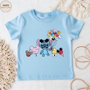 Pu&ograve; includere: T-shirt azzurra con un personaggio dei cartoni animati che tiene in mano palloncini e dolcetti. Il personaggio &egrave; blu con grandi occhi e orecchie nere. La maglietta ha anche un design di zucchero filato, cono gelato e cupcake.