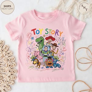 May include: A light pink t-shirt featuring a colorful graphic of characters from the animated film "Toy Story." The design includes the words "Toy Story" in bold letters, along with drawings of Woody, Buzz Lightyear, and other characters.