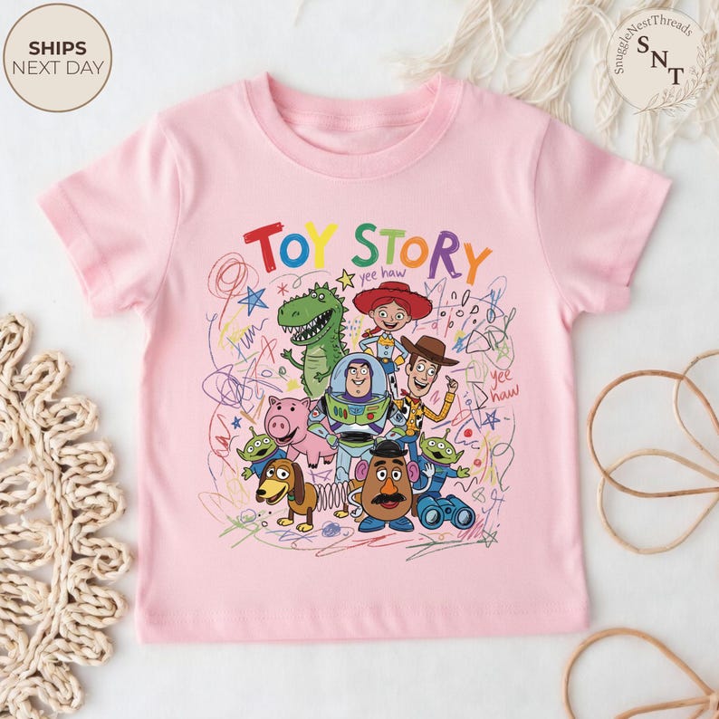 May include: A light pink t-shirt featuring a colorful graphic of characters from the animated film "Toy Story." The design includes the words "Toy Story" in bold letters, along with drawings of Woody, Buzz Lightyear, and other characters.