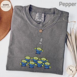 May include: A gray Comfort Colors t-shirt featuring a graphic of green aliens in blue uniforms being lifted by a claw machine. A silver necklace with a pendant is draped on the shirt. The shirt is labeled "Pepper".