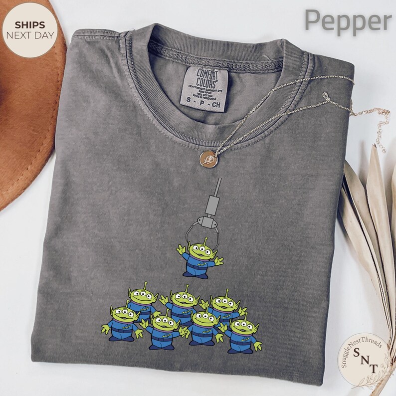 May include: A gray Comfort Colors t-shirt featuring a graphic of green aliens in blue uniforms being lifted by a claw machine. A silver necklace with a pendant is draped on the shirt. The shirt is labeled "Pepper".