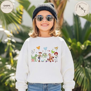 May include: A white sweatshirt featuring a colorful graphic of characters from the animated movie Toy Story. The design includes Woody, Buzz Lightyear, and other characters, along with balloons and stars. The sweatshirt is worn by a child wearing a black beanie and sunglasses.