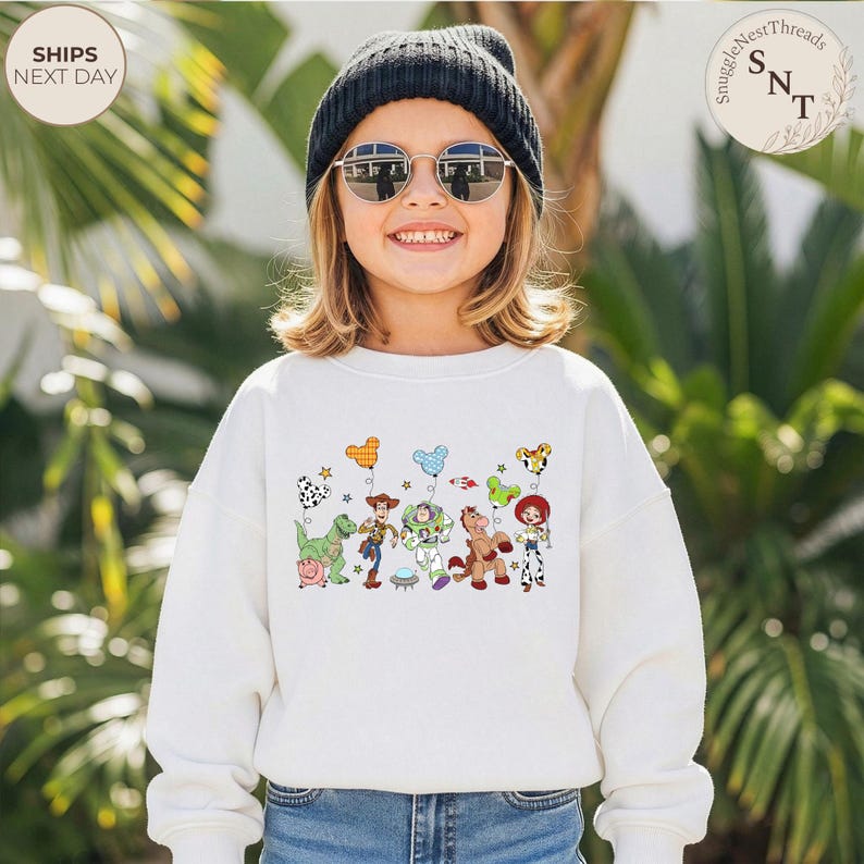 May include: A white sweatshirt featuring a colorful graphic of characters from the animated movie Toy Story. The design includes Woody, Buzz Lightyear, and other characters, along with balloons and stars. The sweatshirt is worn by a child wearing a black beanie and sunglasses.
