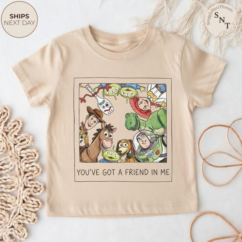 May include: A beige short-sleeved t-shirt featuring a colorful graphic of characters from the movie Toy Story. The graphic is framed and includes Woody, Buzz Lightyear, and other characters. The text "YOU'VE GOT A FRIEND IN ME" is below the graphic.