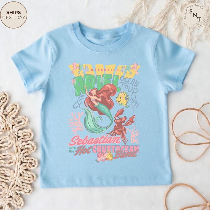 May include: A light blue short-sleeve t-shirt featuring a graphic print of Ariel, Flounder, and Sebastian. The text reads Atlantica Ariel Ocean Tour 1991, Sebastian with the Hot Crustacean Band, accompanied by coral illustrations in pink and yellow.