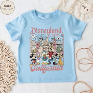 May include: A light blue short-sleeve t-shirt featuring a graphic print of Disneyland California Est. 1955. The design includes illustrations of Mickey Mouse, Minnie Mouse, Donald Duck, Daisy Duck, Goofy, and Pluto in front of the castle.