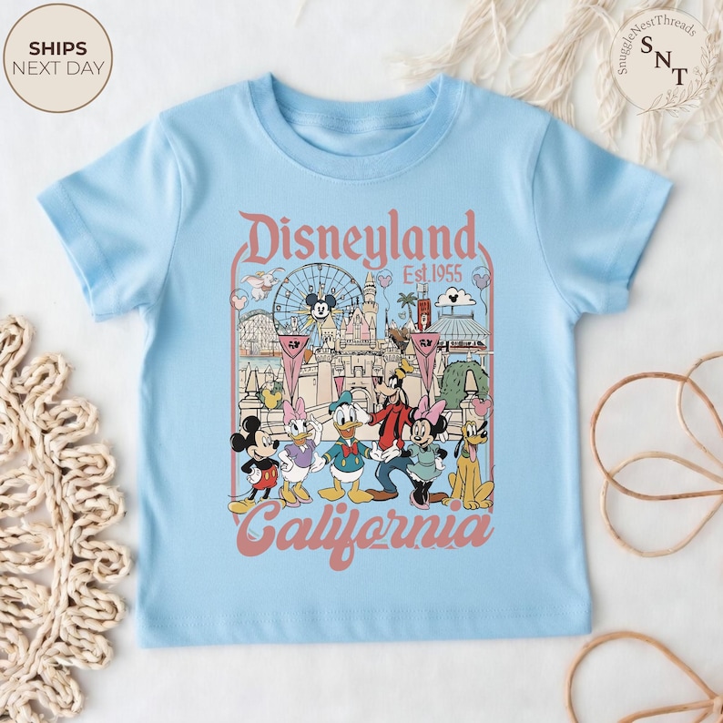 May include: A light blue short-sleeve t-shirt featuring a graphic print of Disneyland California Est. 1955. The design includes illustrations of Mickey Mouse, Minnie Mouse, Donald Duck, Daisy Duck, Goofy, and Pluto in front of the castle.