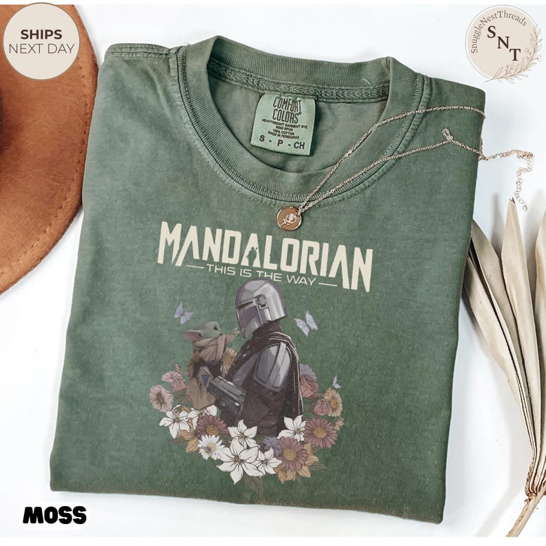 Puede incluir: Una camiseta de cuello redondo color verde musgo con un gr&aacute;fico de The Mandalorian y Grogu rodeados de flores y mariposas. El texto "MANDALORIAN" y "THIS IS THE WAY" aparece en color crema. La camiseta tiene una etiqueta Comfort Colors.