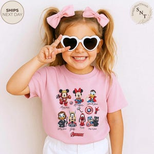 May include: A pink t-shirt featuring various Disney characters, including Mickey Mouse, Minnie Mouse, and Donald Duck. The shirt also displays the Avengers logo. The child is wearing heart-shaped sunglasses and pink bows.