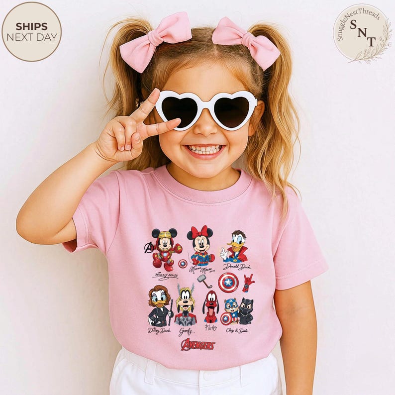 May include: A pink t-shirt featuring various Disney characters, including Mickey Mouse, Minnie Mouse, and Donald Duck. The shirt also displays the Avengers logo. The child is wearing heart-shaped sunglasses and pink bows.
