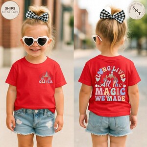 May include: A red t-shirt with the text "OLIVIA" and a castle graphic on the front. The back of the shirt features the text "LONG LIVE All The MAGIC WE MADE". The child is wearing denim shorts and sunglasses.