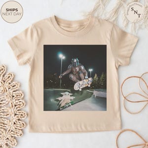 May include: Beige toddler t-shirt featuring a graphic of a person in Mandalorian armor skateboarding, with Grogu on a skateboard below. The image is set in a skate park at night. The shirt has a crew neck and short sleeves.