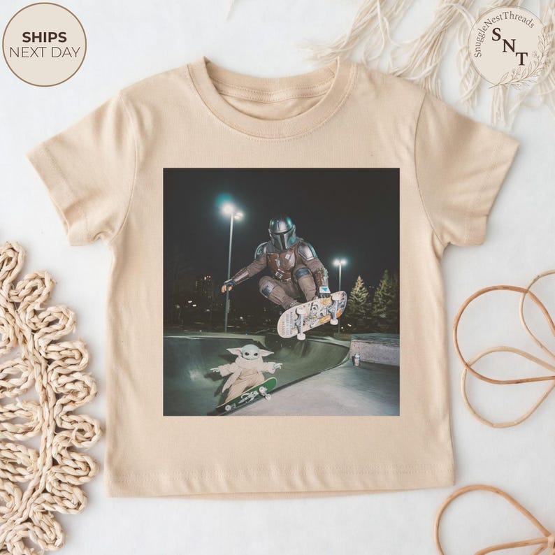 May include: Beige toddler t-shirt featuring a graphic of a person in Mandalorian armor skateboarding, with Grogu on a skateboard below. The image is set in a skate park at night. The shirt has a crew neck and short sleeves.