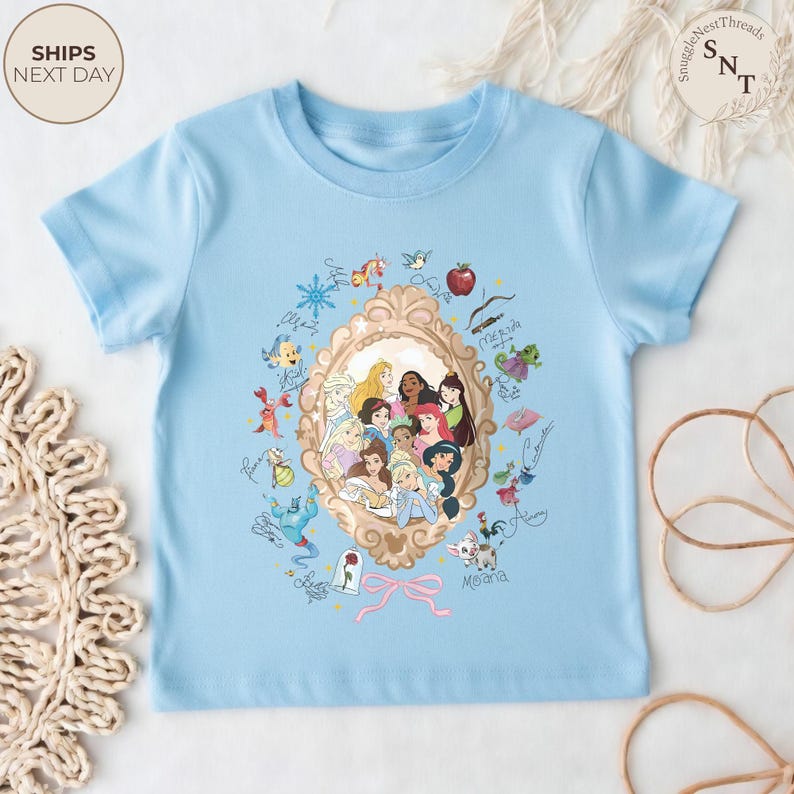 May include: A light blue t-shirt featuring a gold-framed portrait of several animated princesses. Surrounding the portrait are various character illustrations and signatures. The shirt has short sleeves and a crew neck.