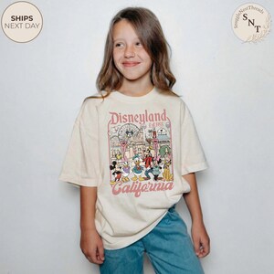 May include: A cream-colored short-sleeve t-shirt featuring a graphic print of Disneyland California with Mickey Mouse, Minnie Mouse, Donald Duck, Daisy Duck, Goofy, and Pluto. The text reads Disneyland Est. 1955 and California in a vintage font.