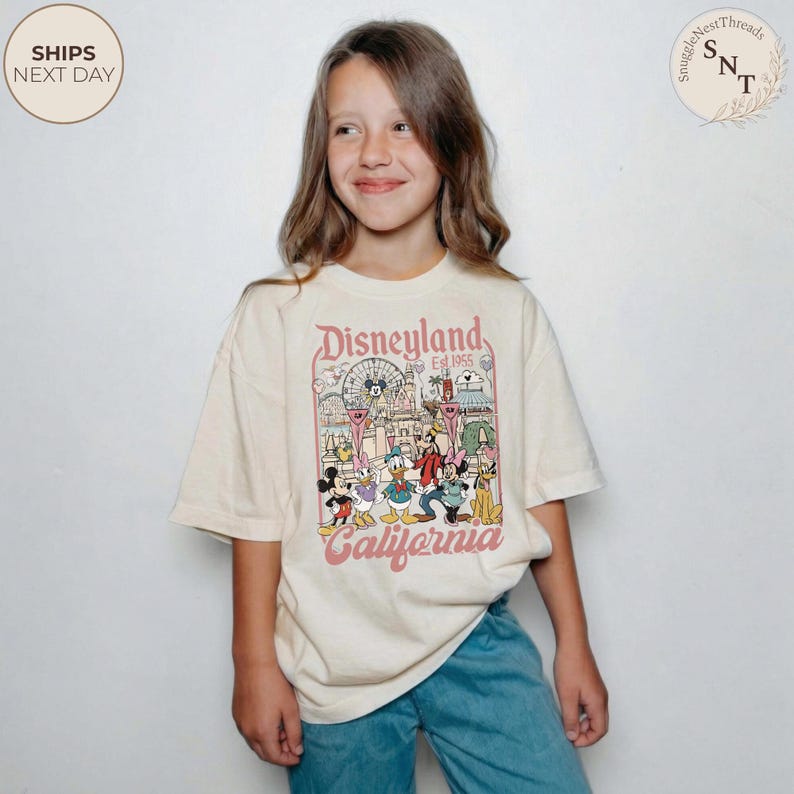 May include: A cream-colored short-sleeve t-shirt featuring a graphic print of Disneyland California with Mickey Mouse, Minnie Mouse, Donald Duck, Daisy Duck, Goofy, and Pluto. The text reads Disneyland Est. 1955 and California in a vintage font.
