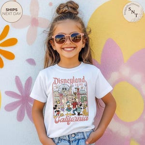 May include: A white short-sleeve t-shirt featuring a colorful graphic print of Disneyland California with the text Disneyland Est 1955 California. The graphic includes illustrations of Mickey Mouse, Minnie Mouse, Donald Duck, Goofy, and Pluto.