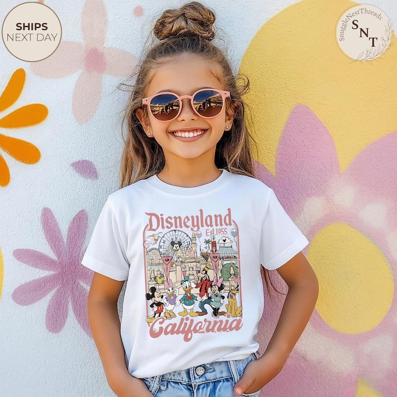 May include: A white short-sleeve t-shirt featuring a colorful graphic print of Disneyland California with the text Disneyland Est 1955 California. The graphic includes illustrations of Mickey Mouse, Minnie Mouse, Donald Duck, Goofy, and Pluto.