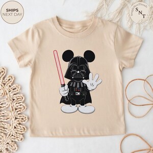 May include: A beige t-shirt featuring a cartoon character mashup of Darth Vader and Mickey Mouse. The character is black with a red lightsaber and white gloves and shoes. The shirt has the text "Ships Next Day".