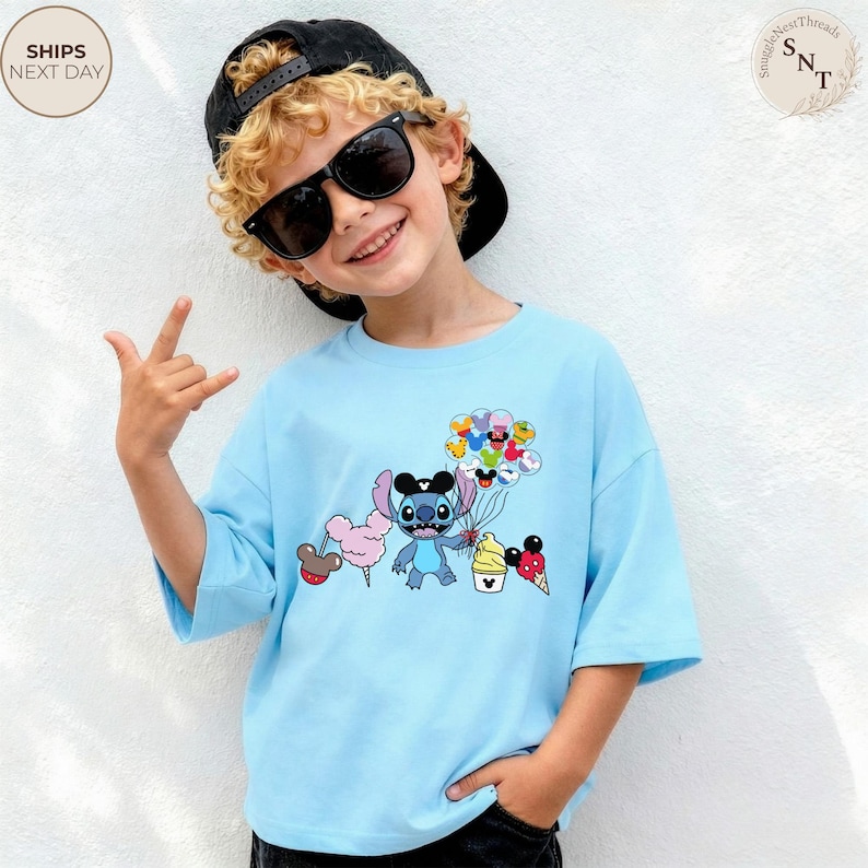 Pu&ograve; includere: T-shirt azzurra con un personaggio dei cartoni animati che tiene in mano palloncini, gelato e zucchero filato. Il personaggio ha grandi orecchie e indossa un cappello nero. La maglietta ha maniche corte e una vestibilit&agrave; ampia. Il testo "SnuggleNestThreads" &egrave; in alto a destra.