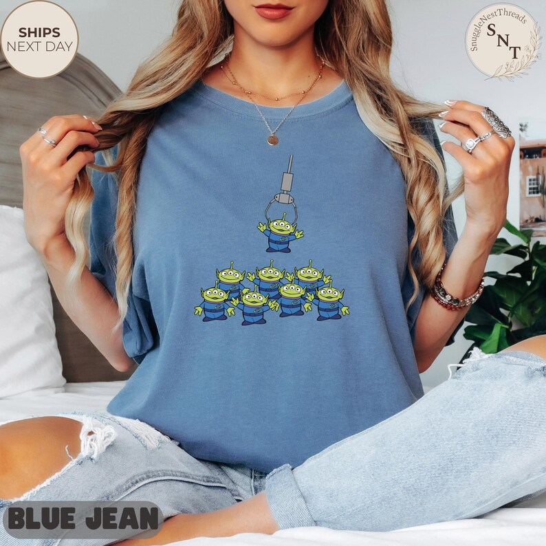 May include: A blue jean colored t-shirt featuring a graphic of the Little Green Men from the Toy Story movie. The design shows a claw machine grabbing one alien, with a group of aliens below. The shirt has the text "BLUE JEAN" at the bottom.