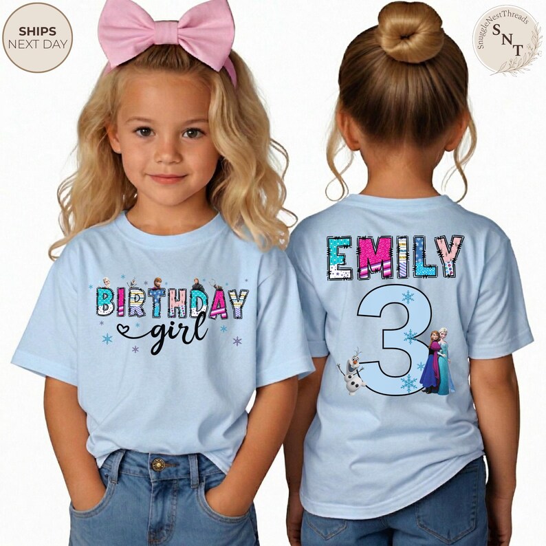 K&ouml;nnte beinhalten: Zwei junge M&auml;dchen tragen hellblaue T-Shirts mit Geburtstagsmotiven. Auf der Vorderseite des Shirts steht "Birthday girl" mit Schneeflocken-Akzenten. Auf der R&uuml;ckseite des Shirts stehen der Name "Emily" und die Zahl "3" mit Frozen-Figuren.