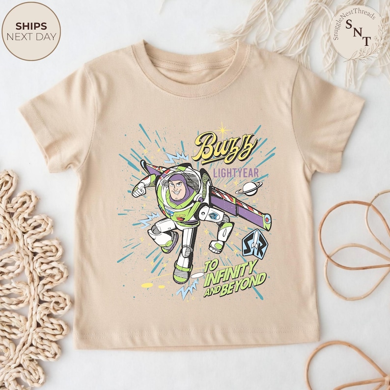 Puede incluir: Una camiseta beige con un gr&aacute;fico de Buzz Lightyear en pose de vuelo, con el texto "Buzz Lightyear" y "To Infinity and Beyond". El dise&ntilde;o incluye un planeta y un logotipo de guardabosques espacial. La camiseta tiene mangas cortas.