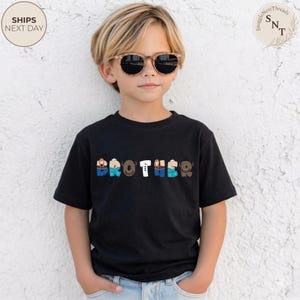 K&ouml;nnte beinhalten: Ein schwarzes T-Shirt mit dem Wort "BROTHER" in bunten, stilisierten Buchstaben. Die Buchstaben sind mit verschiedenen Mustern und Farben gestaltet, darunter Blau, Braun und Wei&szlig;. Das Shirt wird von einem Kind mit Sonnenbrille getragen.