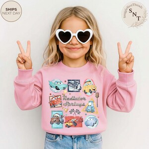 May include: A light pink sweatshirt featuring a colorful graphic of cartoon cars and the text "Radiator Springs". The design includes various car characters in stamp-like frames. The sweatshirt is worn by a child wearing heart-shaped sunglasses.